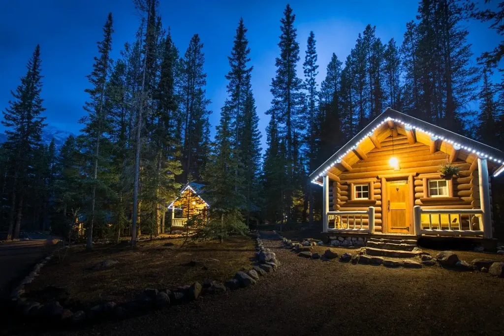 18 Cool 40th Birthday Travel Ideas (with Friends & Family) 18 A cozy log cabin with illuminated eaves sits amid tall pine trees at dusk. Another brightly lit cabin is visible in the background. The scene exudes a serene, rustic charm with the glow of the lights contrasting against the deep blue twilight sky, creating an inviting atmosphere.
