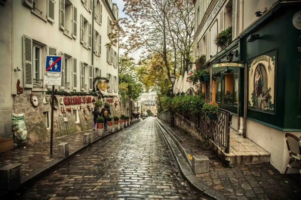 18 Cool 40th Birthday Travel Ideas (with Friends & Family) 14 A narrow, cobblestone street in a quaint European town, reminiscent of those you'll explore on the streets of Paris, flanked by charming buildings with shutters and greenery. The gently sloping street is lined with cozy cafes, small shops, and planters. Trees with autumn leaves add a touch of color to the scene.