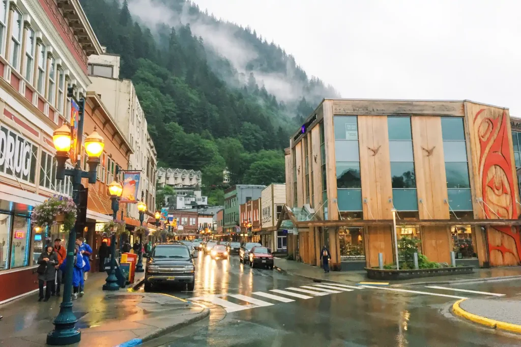 18 Cool 40th Birthday Travel Ideas (with Friends & Family) 7 A rainy street in Juneau, Alaska, features colorful buildings. People with umbrellas walk on sidewalks, while cars drive on wet roads. A mist-covered forested hill rises in the background, and streetlights illuminate the scene.