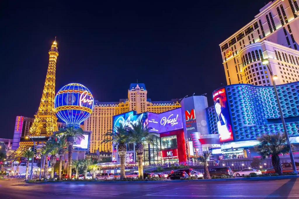 18 Cool 40th Birthday Travel Ideas (with Friends & Family) 11 Night view of the Las Vegas Strip, featuring bright neon lights and prominent hotel-casinos. The Eiffel Tower replica at Paris Las Vegas shines beside vibrant signs, including the Planet Hollywood and MGM Grand marquees. Palm trees line the bustling street, epitomizing the essence of Las Vegas.