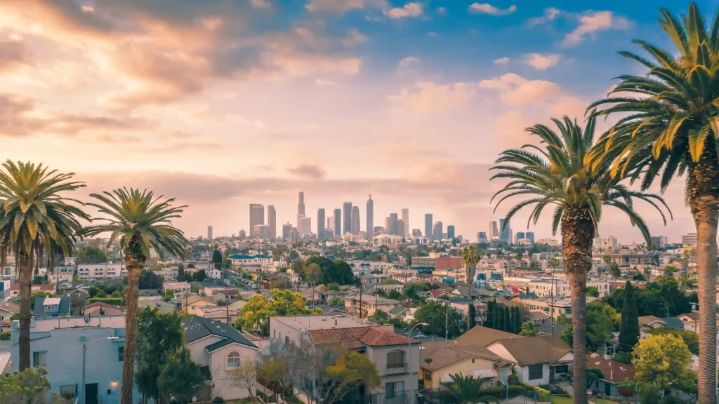 18 Cool 40th Birthday Travel Ideas (with Friends & Family) 4 Skyline of Los Angeles with tall buildings in the distance, framed by palm trees. The foreground features residential houses with red-tiled roofs, and the sky above is a mix of soft clouds and pastel hues from the setting or rising California sun.