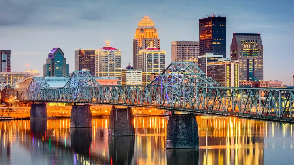 18 Cool 40th Birthday Travel Ideas (with Friends & Family) 3 A city skyline at dusk featuring Louisville's prominent, illuminated bridge in the foreground with bright lights reflecting on the water below. Skyscrapers and buildings of various heights, some with colorful lighting, dominate the background under a cloudy evening sky.