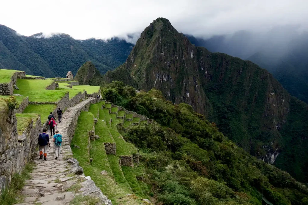 18 Cool 40th Birthday Travel Ideas (with Friends & Family) 16 A group of people hike along a stone path surrounded by lush green terraces, reminiscent of Machu Picchu Hiking. In the background, a towering, jagged peak rises into a foggy, cloud-covered sky. Dense forested hills form a dramatic backdrop, creating a serene and adventurous scene