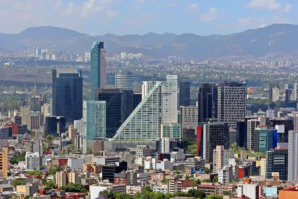 18 Cool 40th Birthday Travel Ideas (with Friends & Family) 10 Aerial view of Mexico City's skyline with various high-rise buildings and skyscrapers in the foreground. The landscape includes modern glass buildings, residential areas, and lush greenery. Mountains and a partly cloudy sky are visible in the background.