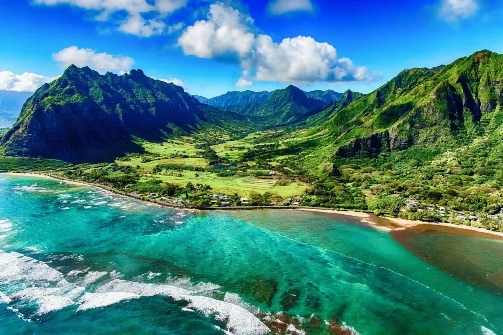 18 Cool 40th Birthday Travel Ideas (with Friends & Family) 6 A stunning aerial view of a lush, green valley in Oahu, Hawaii, surrounded by dramatic mountain ranges. The clear, turquoise ocean meets the shore, with white waves breaking along the sandy beach. The landscape is vibrant under a bright blue sky dotted with fluffy white clouds.