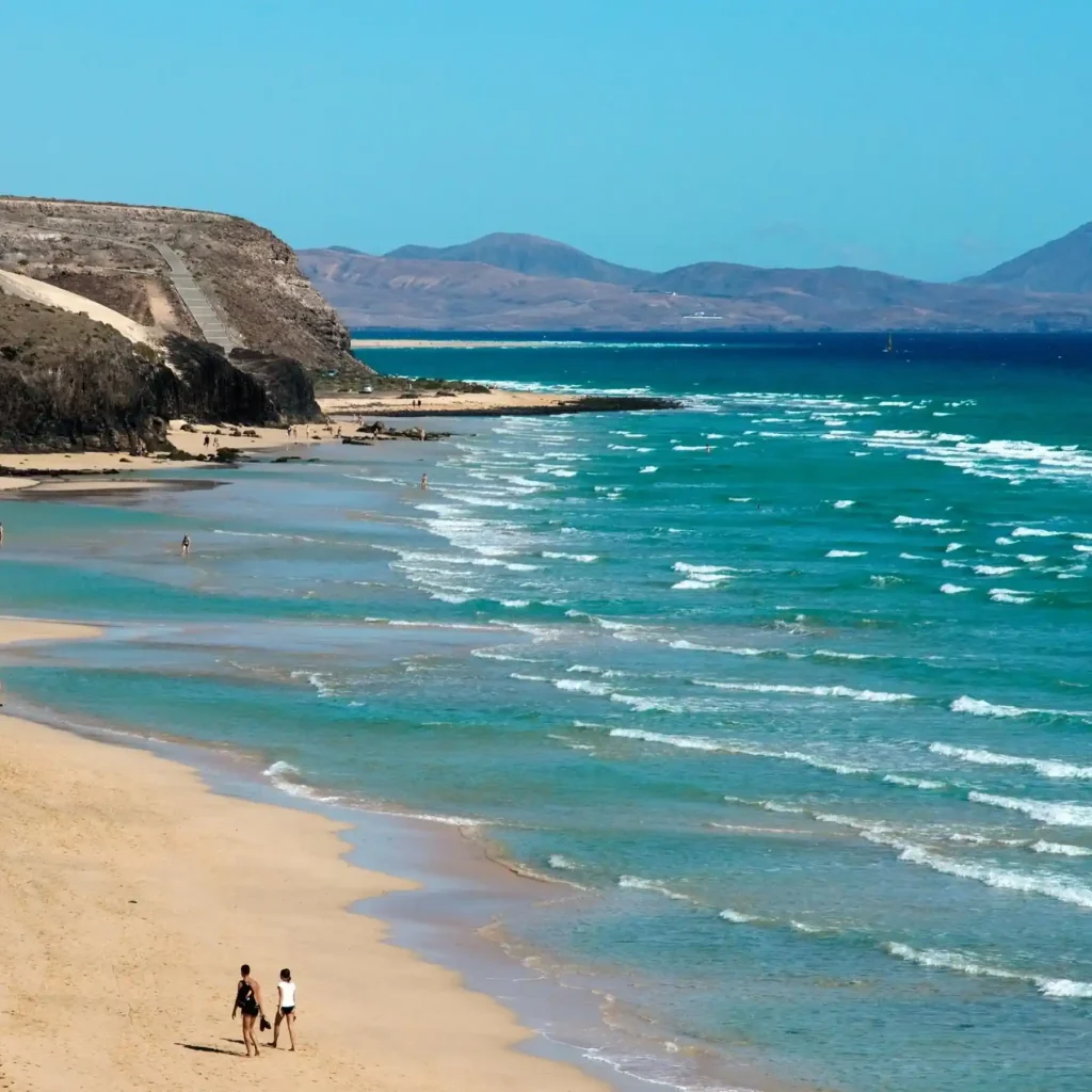 15 Beaches on Earth That Don't Feel Real, According to YouTube 2 A serene beach scene at Sotavento Beach with a golden sandy shore gently curving along the coastline. A few people are walking and relaxing near the water. The turquoise ocean waves calmly lap against the shore. Rocky landscapes and distant mountains frame the background under a clear blue sky.