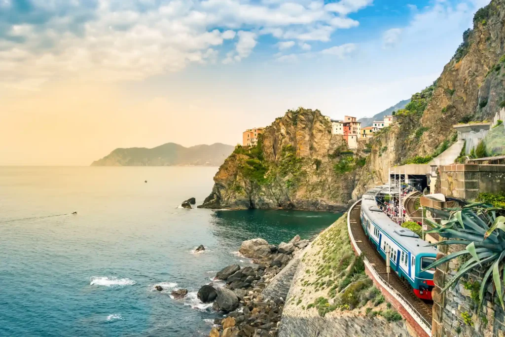 18 Cool 40th Birthday Travel Ideas (with Friends & Family) 17 A scenic coastal train travels along a rocky cliffside with the turquoise sea on the left. Colorful buildings are perched on the cliff with mountains in the background under a partly cloudy sky. This picturesque scene showcases the beauty of Italy's coastline as the train moves towards the horizon.