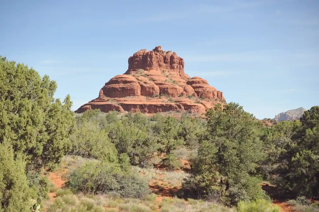 12 Easy Hikes in Sedona AZ with Stunning Red Rock Views 3 A striking red rock formation rises against a clear blue sky along the Bell Rock Trail, flanked by green desert shrubs and trees. The rock's layered, dome-like appearance contrasts with the surrounding greenery, creating a scenic desert landscape.