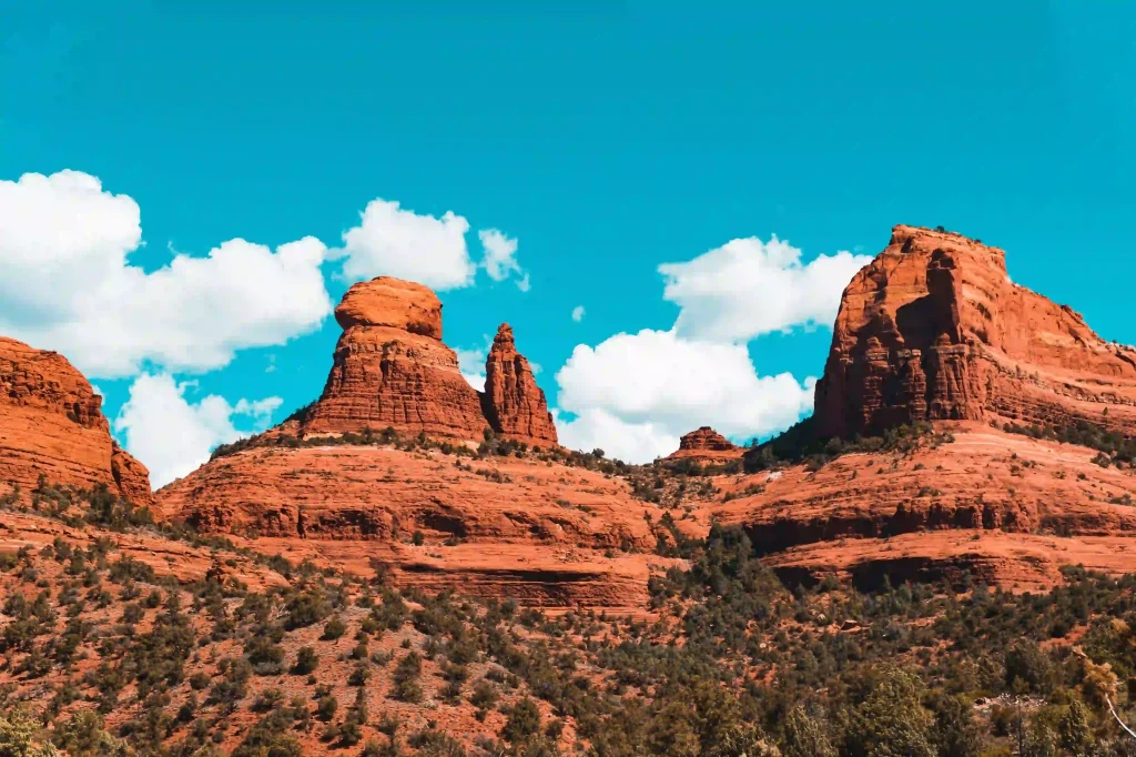 12 Easy Hikes in Sedona AZ with Stunning Red Rock Views 10 A picturesque view of Sedona's red rock formations under a bright blue sky dotted with fluffy white clouds greets hikers on the Big Park Loop. The rugged terrain, featuring prominent rock structures and scattered green vegetation, creates a stunning contrast against the clear daytime sky.