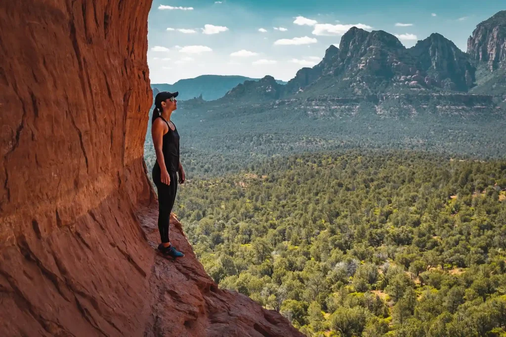 12 Easy Hikes in Sedona AZ with Stunning Red Rock Views 4 A person in athletic wear stands on a rocky ledge, looking out over a vast, forested valley with a backdrop of rugged mountains beneath a partly cloudy sky. The scene captures a sense of adventure and natural beauty near the renowned Birthing Cave.