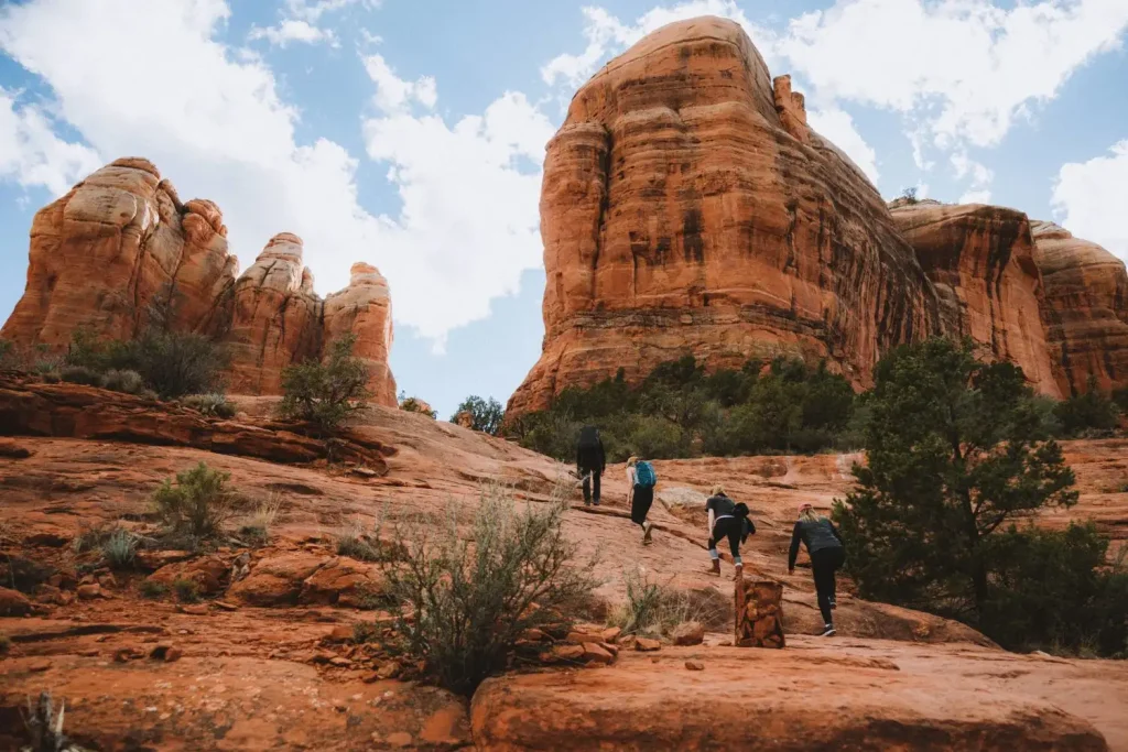 12 Easy Hikes in Sedona AZ with Stunning Red Rock Views 12 A group of people hiking up the rocky, red sandstone Cathedral Rock Trail, surrounded by towering rock formations and sparse vegetation. The sky is partly cloudy.