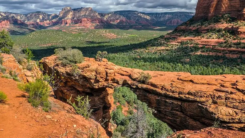 12 Easy Hikes in Sedona AZ with Stunning Red Rock Views 6 The image shows an expansive view along the Devil's Bridge Trail in Sedona, Arizona. The large sandstone arch stretches across a deep ravine with vibrant red and green vegetation below. Rugged cliffs and forested areas extend into the distance under a partly cloudy sky.