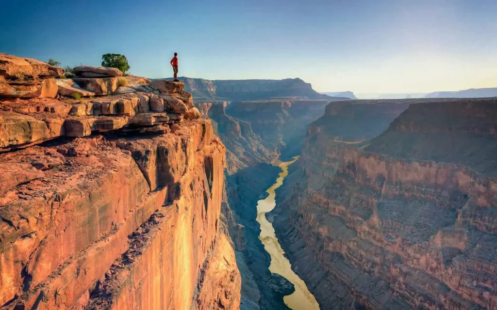 10 Best Spring Break Destinations for Families on a Budget in US 5 A person stands at the edge of a high cliff, gazing over the vast, winding Grand Canyon below. The scene features steep, rocky walls and a river flowing through the bottom of the canyon in Arizona, with a bright, clear sky above.