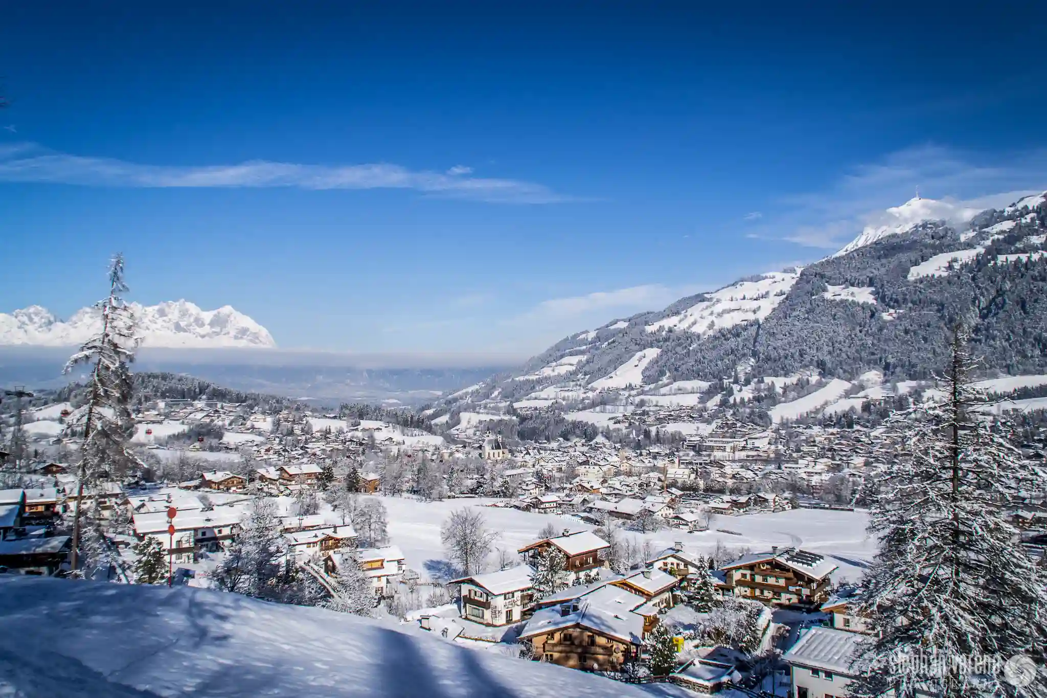10 Best European Countries to Visit in December This Year 7 A snowy winter landscape featuring a village with snow-covered rooftops nestled in a valley. Surrounding hills and mountains, reminiscent of Kitzbühel in Austria, are blanketed with snow, and the sky is clear blue, offering a picturesque view. Trees and houses are scattered throughout the scene.