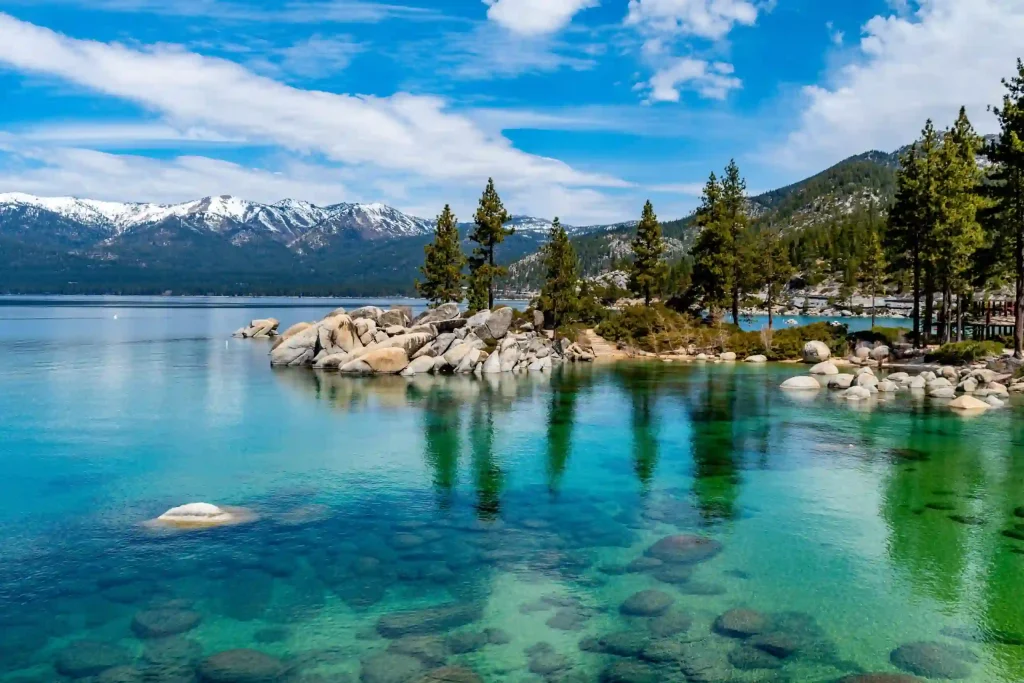 10 Best Spring Break Destinations for Families on a Budget in US 10 Lake Tahoe boasts a serene expanse of crystal-clear turquoise water with visible rocks beneath the surface. Towering pine trees stand on rocky outcrops by the lakeside, while snow-capped mountains rise majestically in the background under a partly cloudy Nevada sky.