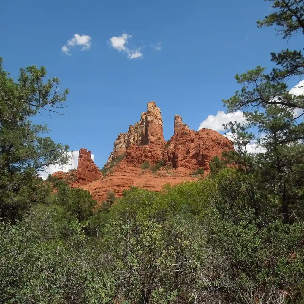 12 Easy Hikes in Sedona AZ with Stunning Red Rock Views 9 Rock formations in Sedona, Arizona, rise against a blue sky with a few clouds. The rugged, red-hued cliffs are surrounded by green trees and shrubs along Margs Draw Trail, creating a vivid contrast. The natural landscape exudes a sense of tranquility and beauty.