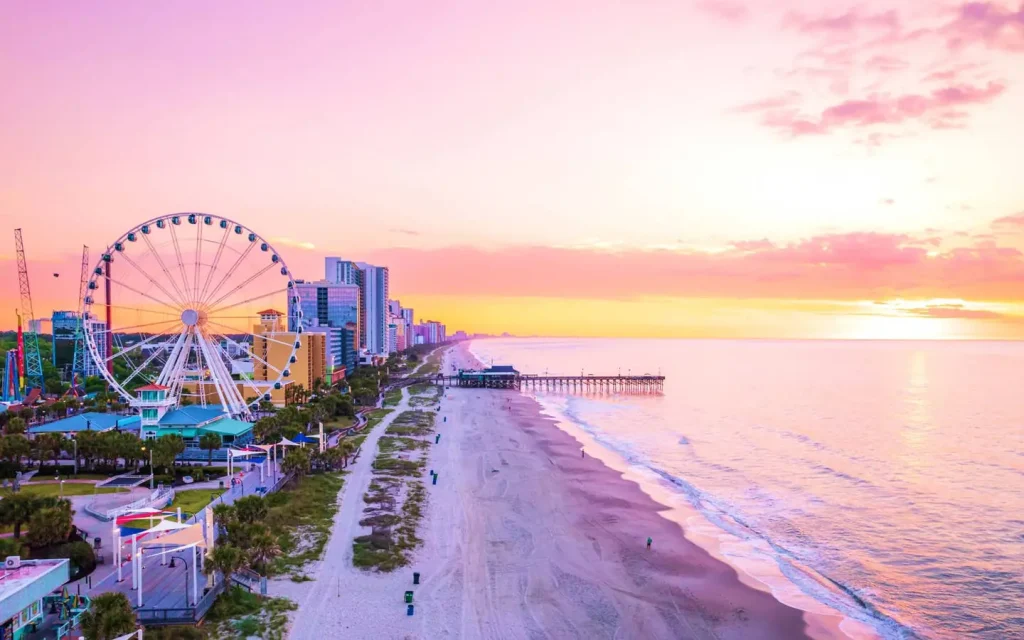 10 Best Spring Break Destinations for Families on a Budget in US 1 A coastal cityscape at sunset in Myrtle Beach, South Carolina, with a Ferris wheel, tall buildings, and a pier extending into the ocean. The sky is pink and orange, and the beach is mostly empty with a few people strolling along the shore.