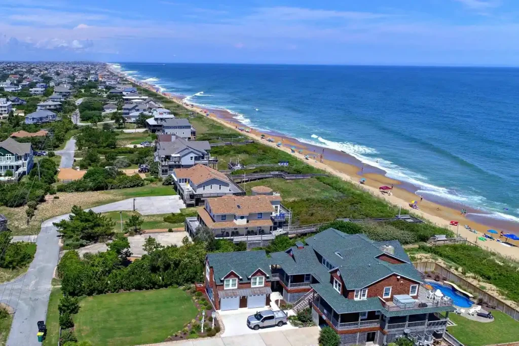 10 Best Spring Break Destinations for Families on a Budget in US 4 An aerial view of a coastal town in North Carolina's Outer Banks shows houses lining the shore. The beach has a few sunbathers and umbrellas, while waves gently crash onto the sandy shore. Greenery surrounds the residential area, and the ocean stretches to the horizon under a blue sky with few clouds.