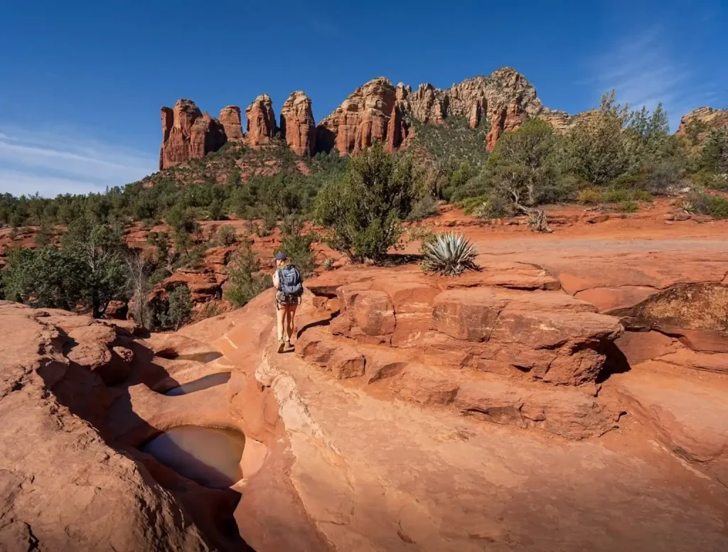 12 Easy Hikes in Sedona AZ with Stunning Red Rock Views 8 A hiker wearing a backpack walks on the rugged, rocky terrain of Soldier Pass Trail with small pools of water. Tall, red rock formations and lush green vegetation surround the area. A clear blue sky stretches above the scenic landscape.