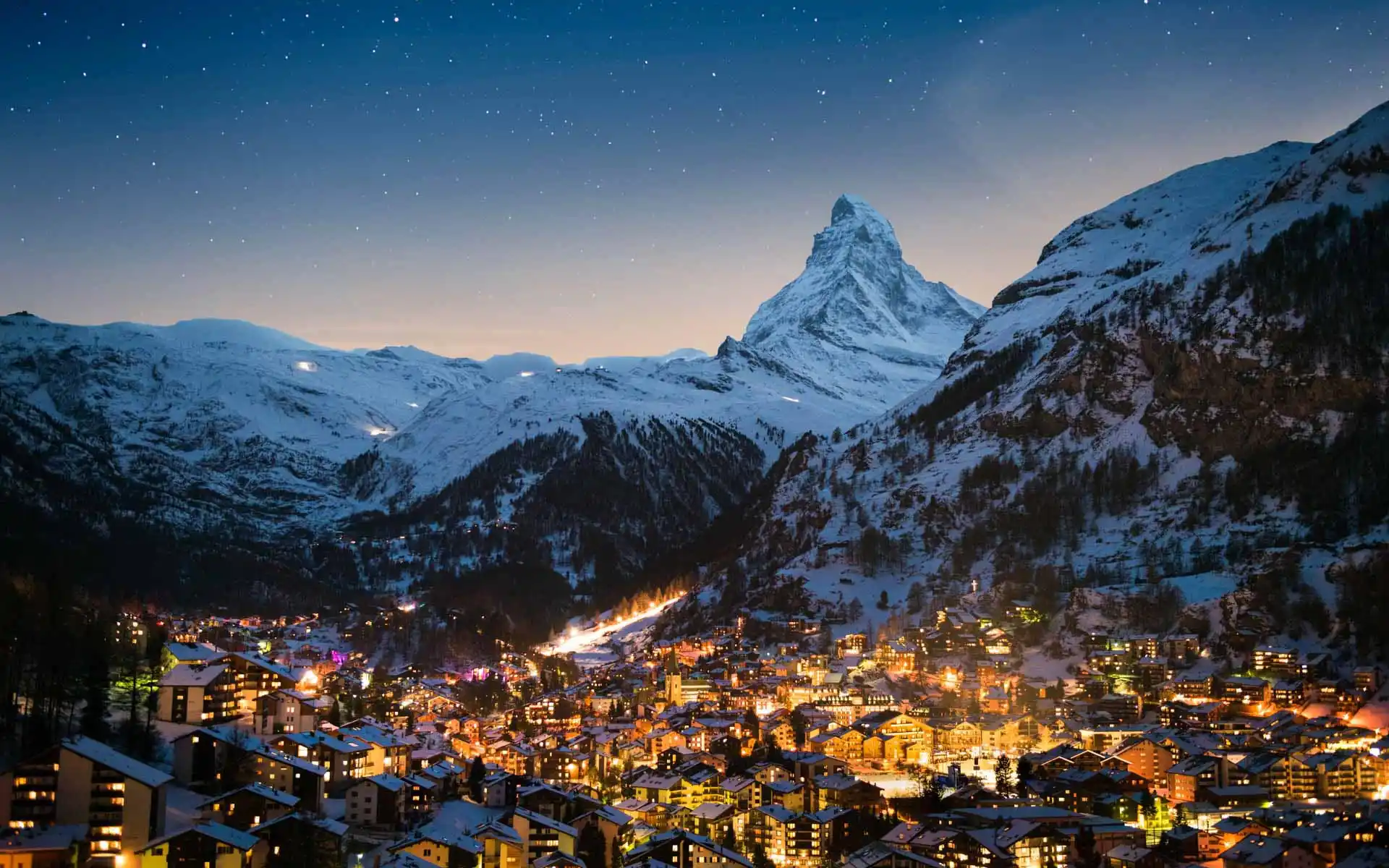 10 Best European Countries to Visit in December This Year 8 A nighttime view of a snow-covered mountainous region in Zermatt, Switzerland, with a prominent, pyramid-shaped peak in the background. Below, a village illuminated with warm lights nestles in a valley, surrounded by snow-dusted buildings and trees. Stars dot the clear sky above.