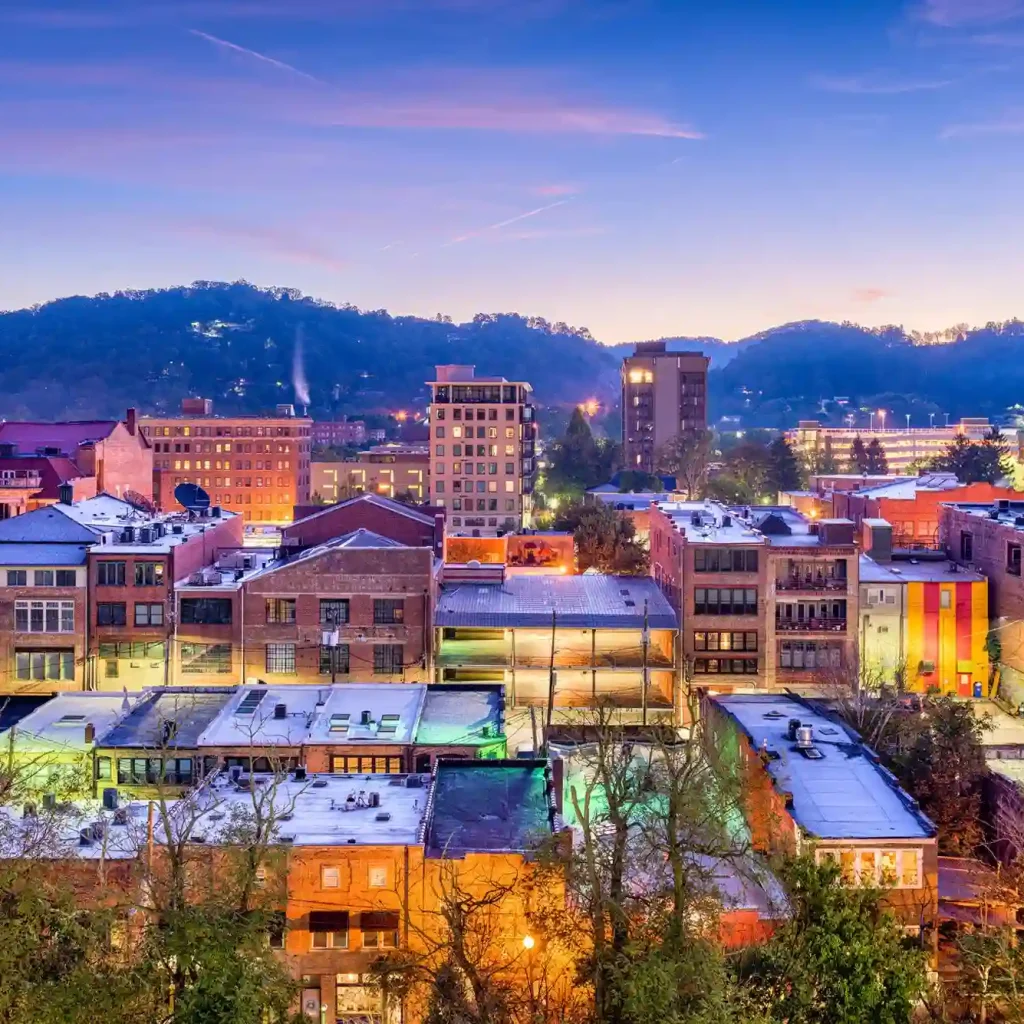 Long Weekend Vacations East Coast: 12 Budget-Friendly Places You’ll Love 5 A vibrant Asheville cityscape at dusk features colorful buildings in the foreground, framed by tree-covered hills typical of North Carolina. The sky is a gradient of blue and orange, adding to the scenic view.