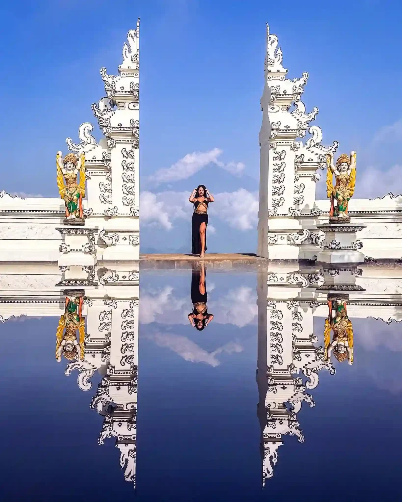 12 Best Christmas Beach Holiday Destinations Around the World 11 A woman stands between two intricately carved white temple gates in Bali, mirrored by a reflective surface below. She poses against a bright blue sky with clouds, flanked by two vibrant guardian statues. The scene creates a symmetrical, ethereal effect.