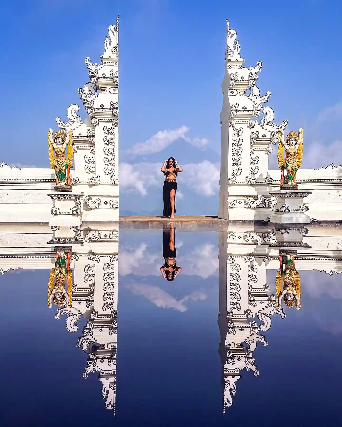 20 Bucket-List Worthy Exotic Travel Destinations to Visit Now 2 A woman stands between two intricately carved white temple gates in Bali, mirrored by a reflective surface below. She poses against a bright blue sky with clouds, flanked by two vibrant guardian statues. The scene creates a symmetrical, ethereal effect.