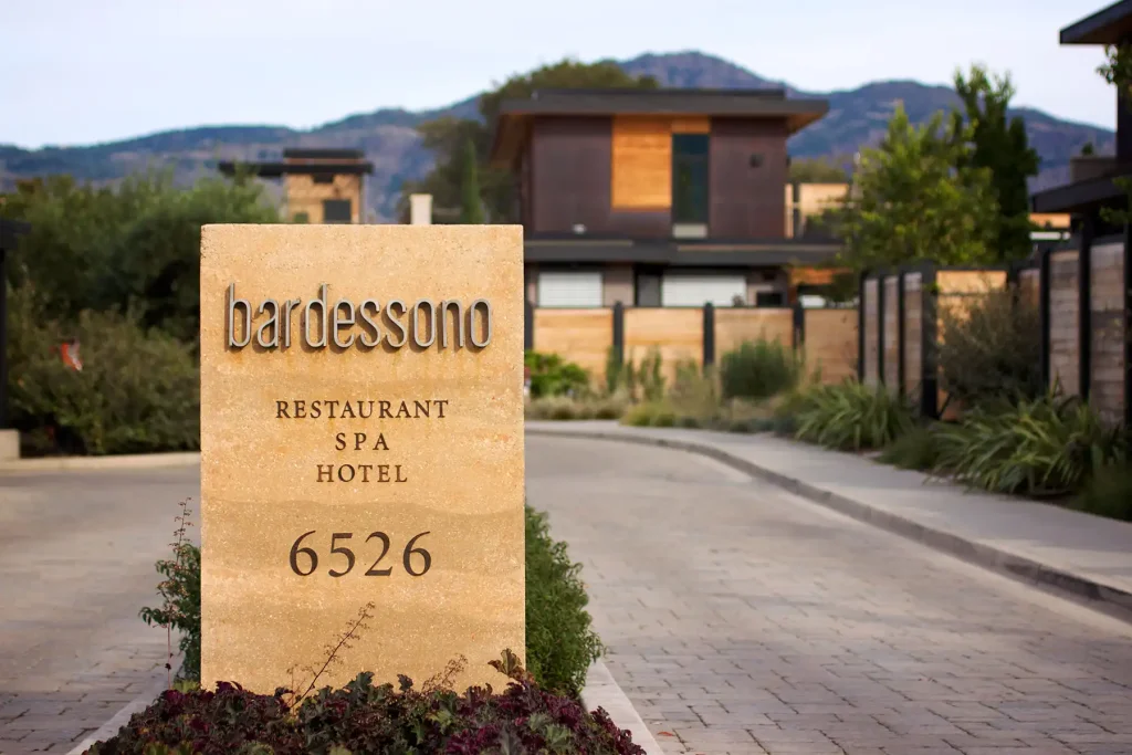 Sign for Bardessono Hotel and Spa with "RESTAURANT," "SPA," "HOTEL," and "6526" displayed. Background shows a modern building and a mountain.
