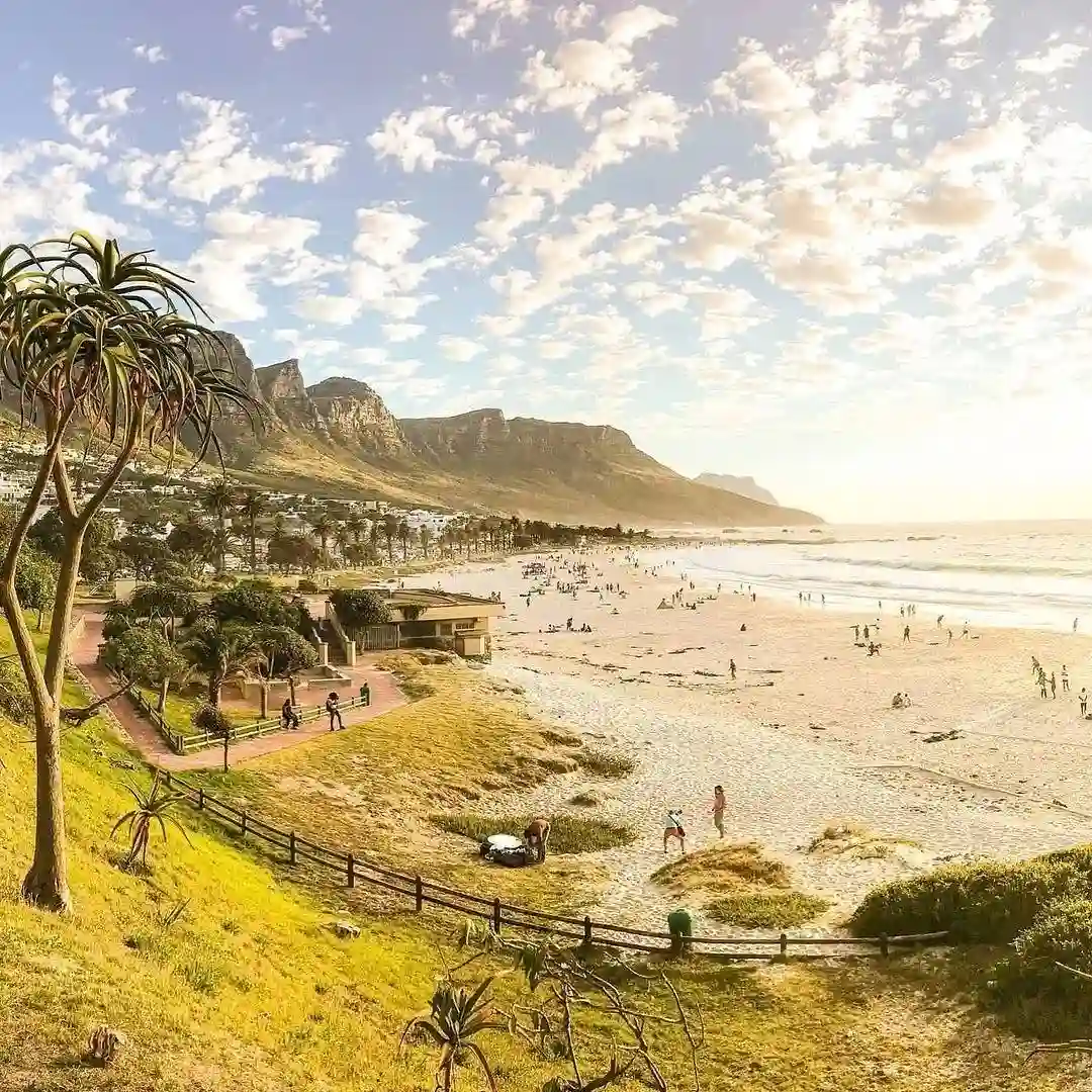 20 Bucket-List Worthy Exotic Travel Destinations to Visit Now 8 A scenic beach view with a golden sunset, soft waves, and scattered beachgoers. Mountains rise in the background, and a few trees and buildings line the coast. A wooden fence winds along a grassy hill overlooking the beach in Cape Town. The sky is dotted with clouds.