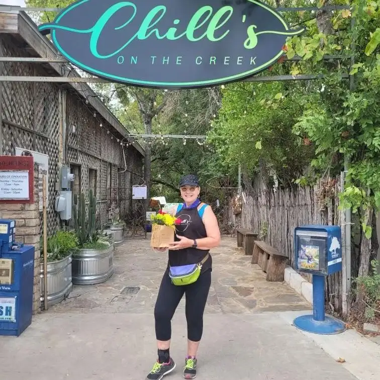 The Romantic Guide to Spoon Mountain Glamping for Beginners 12 A person stands smiling outside Chill's On The Creek, holding a brown paper bag filled with vibrant flowers.