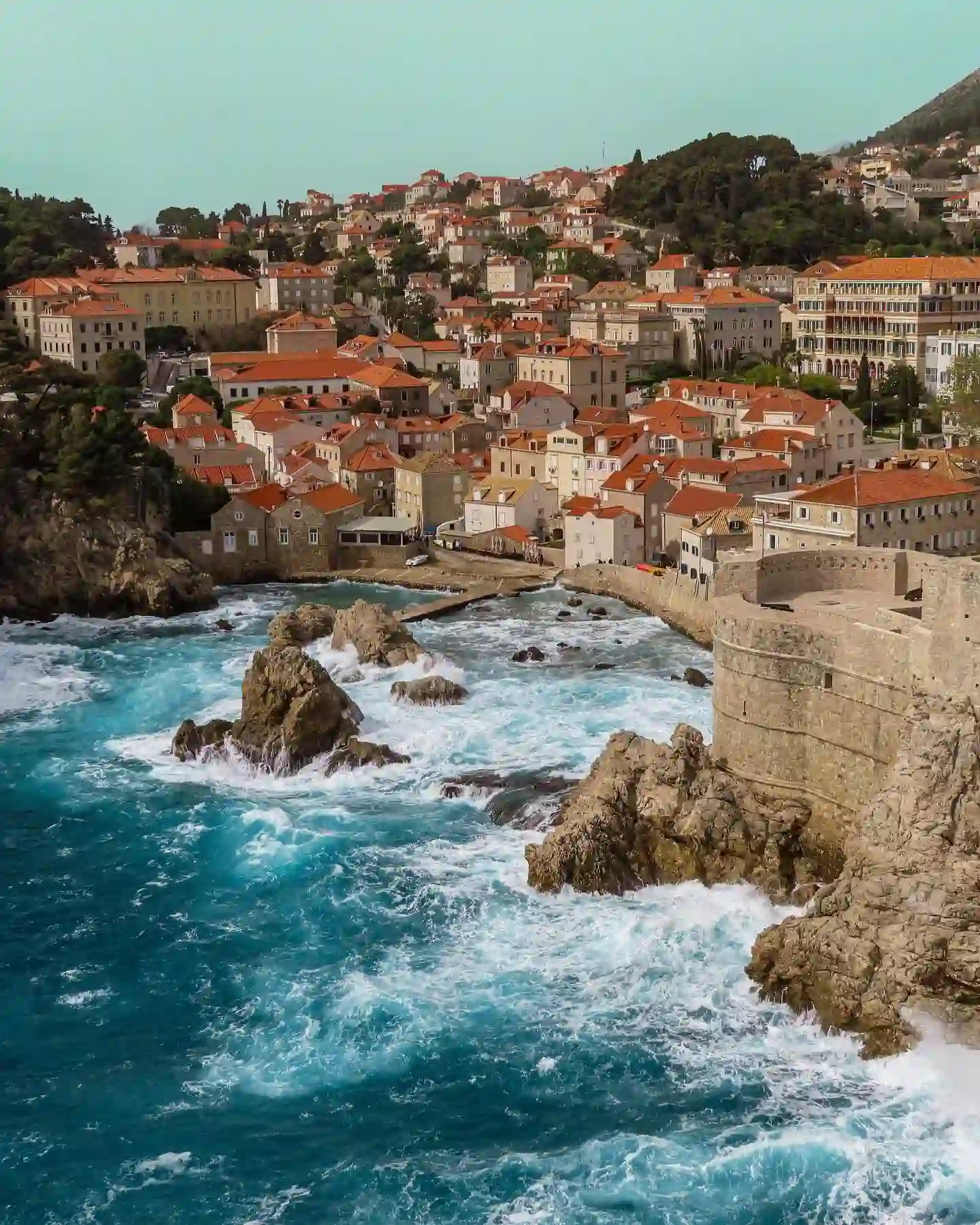 20 Bucket-List Worthy Exotic Travel Destinations to Visit Now 16 A coastal town with numerous orange-roofed buildings is seen against a backdrop of green hills. Situated along a rocky coastline in Croatia, with waves crashing against the shore, Dubrovnik's stone fortification extends into the water, adding to the picturesque scenery.