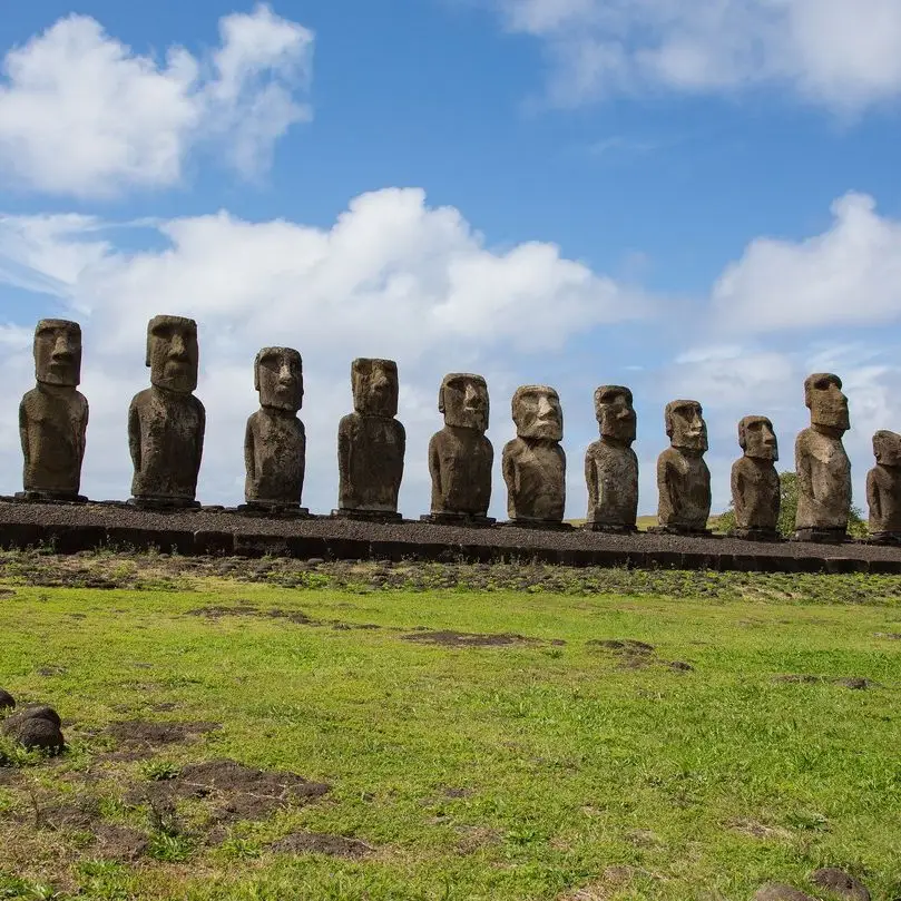 20 Bucket-List Worthy Exotic Travel Destinations to Visit Now 15 A line of Moai statues stands on a stone platform under a partly cloudy sky on Easter Island, Chile. The statues, carved from volcanic tuff, face inland, displaying stoic expressions. The foreground features grassy terrain with scattered rocks.