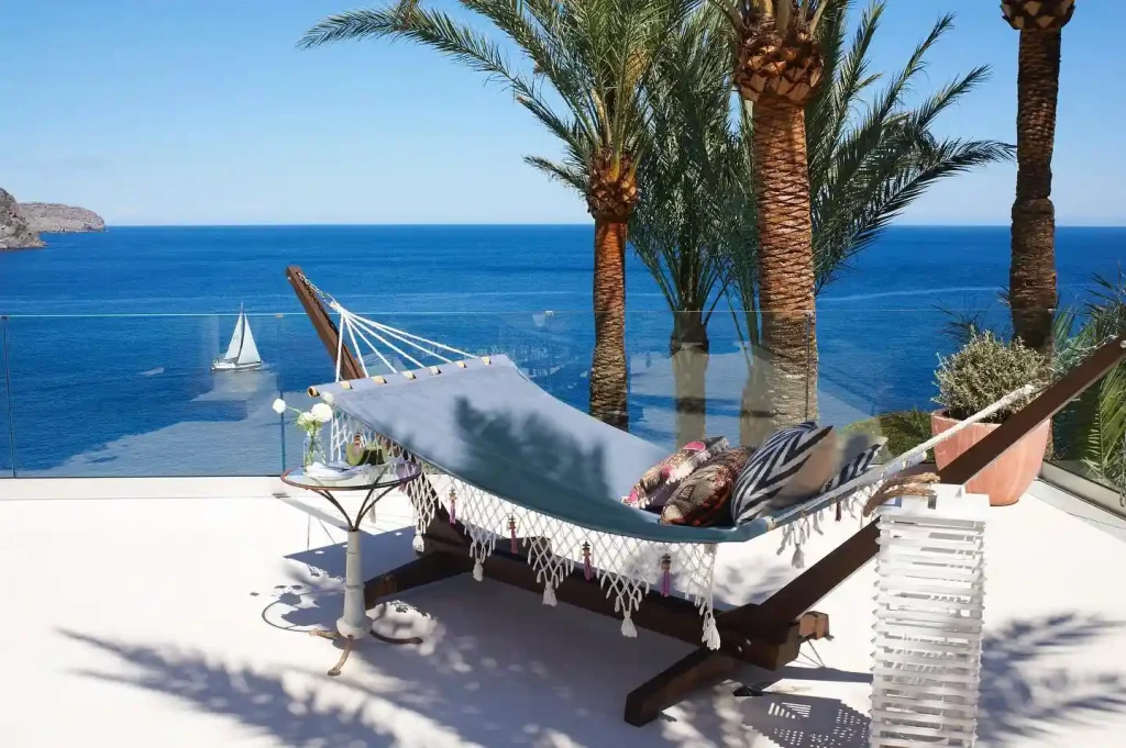 20 Bucket-List Worthy Exotic Travel Destinations to Visit Now 20 A hammock with decorative pillows is set up on a white patio overlooking the clear blue ocean of Ibiza. Palm trees provide shade, and a sailboat is visible in the distance on the water. A glass railing borders the patio, offering an unobstructed view of this serene seascape in Spain.