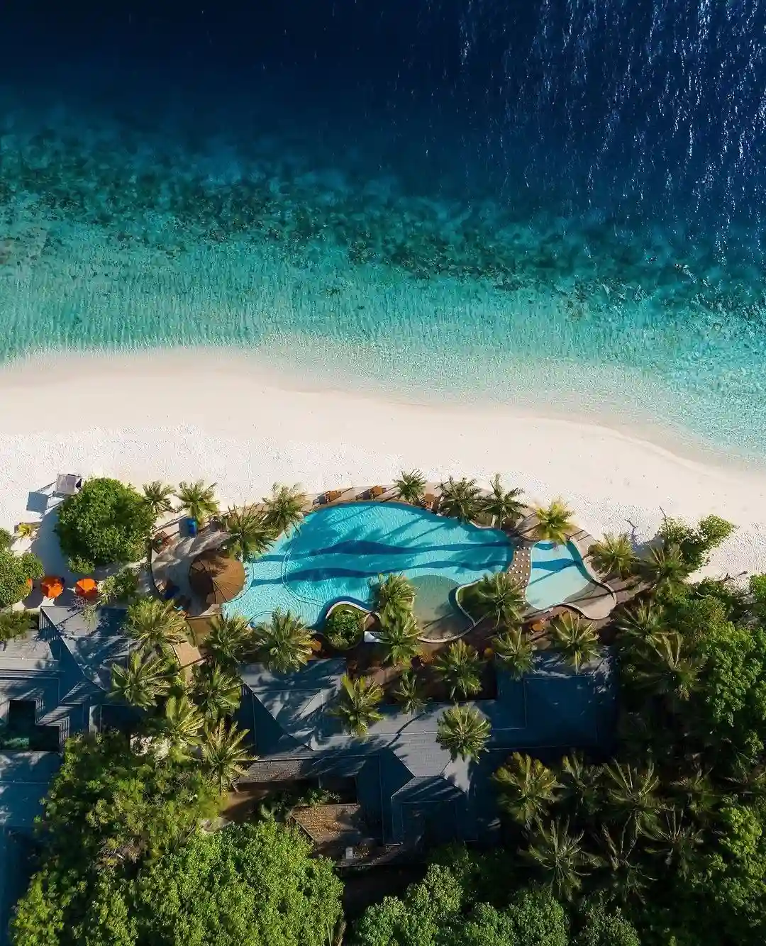 20 Bucket-List Worthy Exotic Travel Destinations to Visit Now 1 Aerial view of a tropical beach in the Maldives with a turquoise swimming pool surrounded by palm trees and lush greenery. The pool area is adjacent to white sand leading to a calm, clear blue ocean.