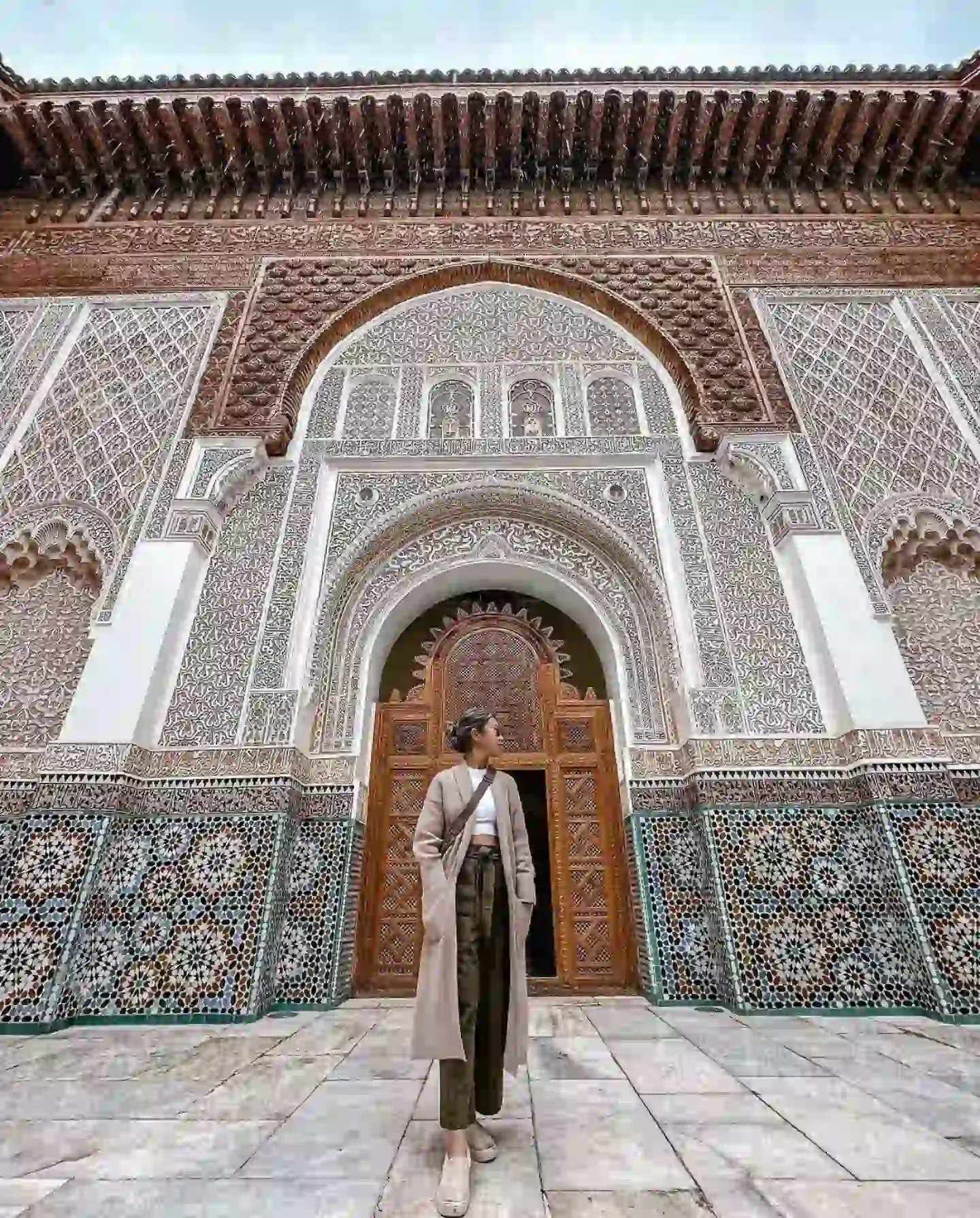 20 Bucket-List Worthy Exotic Travel Destinations to Visit Now 3 A person in a beige coat and hat stands in front of an intricately designed building with arches and ornate tile work, reminiscent of Marrakesh. The architecture features detailed geometric patterns and traditional motifs. The large wooden doors add a rich, cultural element to the scene.