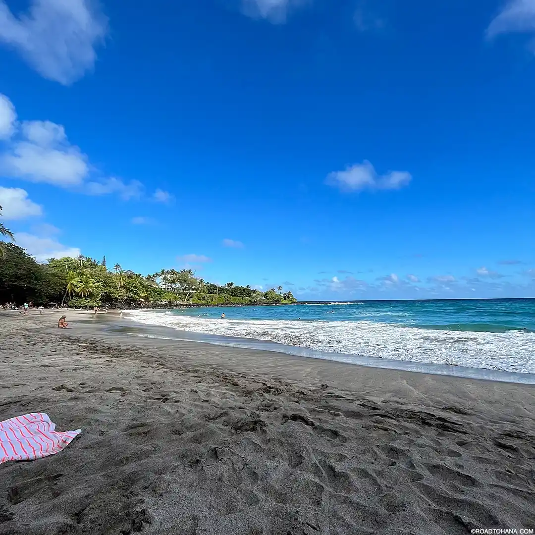 20 Bucket-List Worthy Exotic Travel Destinations to Visit Now 17 A serene beach in Maui with dark sand, clear blue water, and a slightly cloudy sky. There are a few people in the distance enjoying the beach, and a striped towel is laid near the foreground. Lush greenery, quintessential of Hawaii, is visible in the background.