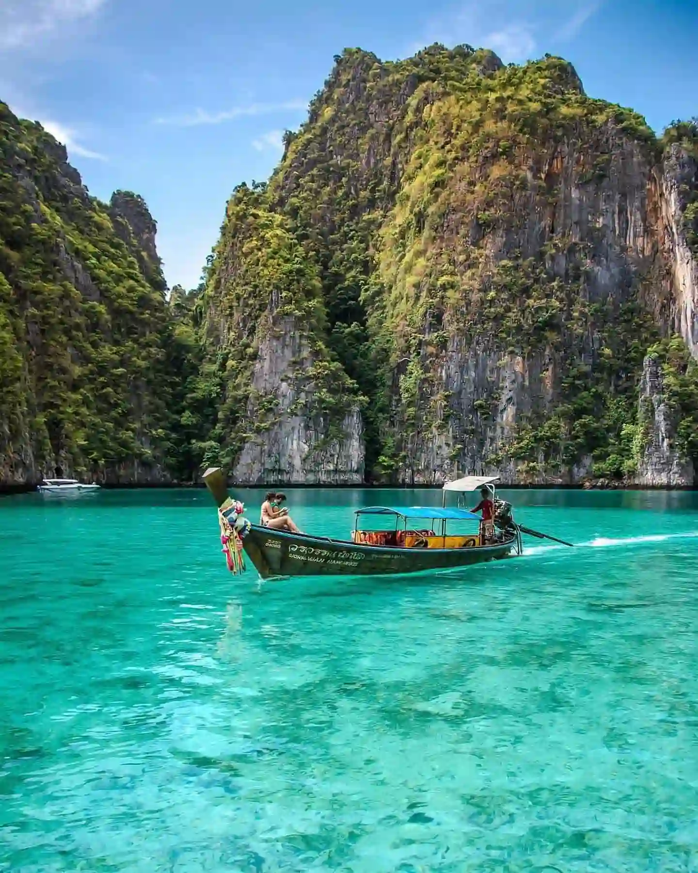 20 Bucket-List Worthy Exotic Travel Destinations to Visit Now 12 A traditional wooden long-tail boat with passengers moves through clear turquoise waters surrounded by towering limestone cliffs covered in lush greenery, under a bright blue sky. The scene, reminiscent of Phuket's picturesque tropical landscape in Thailand, depicts tranquility at its finest.