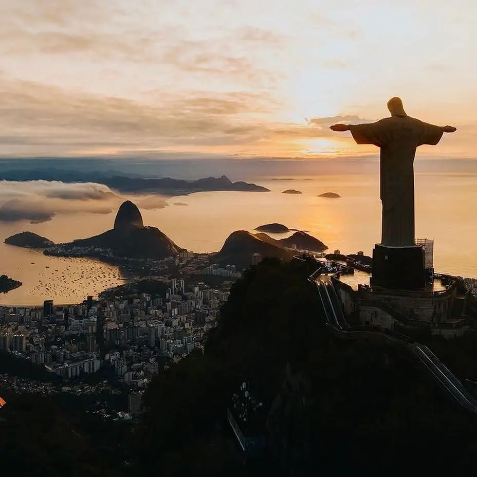 20 Bucket-List Worthy Exotic Travel Destinations to Visit Now 6 A breathtaking view at sunset of the Christ the Redeemer statue atop Corcovado Mountain in Rio de Janeiro, Brazil. The sprawling cityscape and Sugarloaf Mountain are visible below, with the glowing sun setting over the ocean.