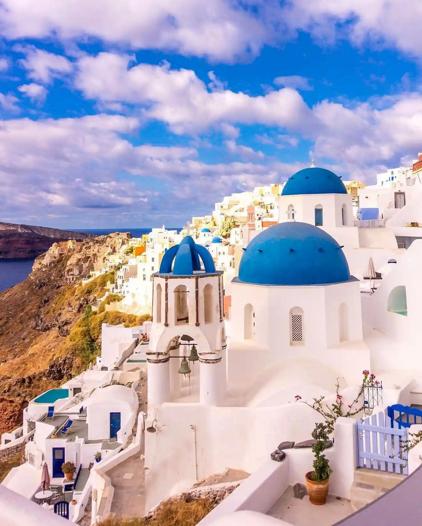 20 Bucket-List Worthy Exotic Travel Destinations to Visit Now 4 A picturesque view of Santorini, Greece, showcasing its white-washed buildings with iconic blue domes perched on a hillside above the deep blue Aegean Sea. The clear sky is dotted with a few clouds, and vibrant colors adorn the scene, creating a serene atmosphere in Santorini's enchanting landscape.