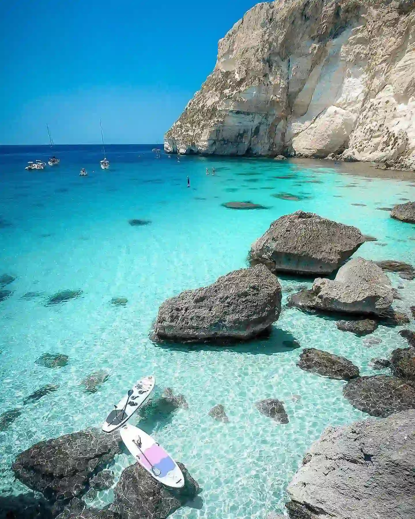 20 Bucket-List Worthy Exotic Travel Destinations to Visit Now 19 A stunning coastal scene in Sardinia features clear turquoise waters with rocky cliffs in the background. Several boats are anchored in the bay, and two paddleboards rest on the calm, shallow water near large rocks. The sky is clear and bright blue.