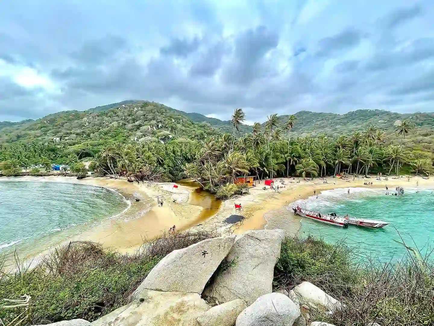 20 Bucket-List Worthy Exotic Travel Destinations to Visit Now 14 A scenic view of a tropical beach with golden sand and lush green hills in the background. The shoreline curves around a small bay, meeting clear turquoise waters. Boats are docked near the shore, and palm trees line the beach, providing shade to visitors in Tayrona National Park, Colombia.