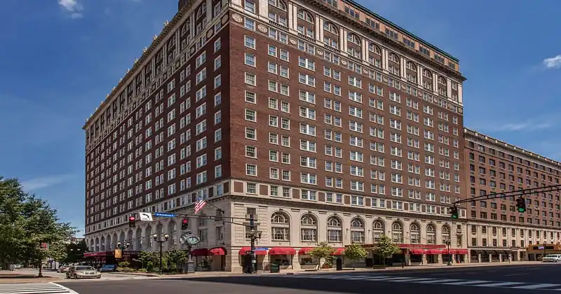 A large, multi-story brick building with numerous windows, flags, and awnings stands on a corner street under a clear blue sky. This is The Brown Hotel.