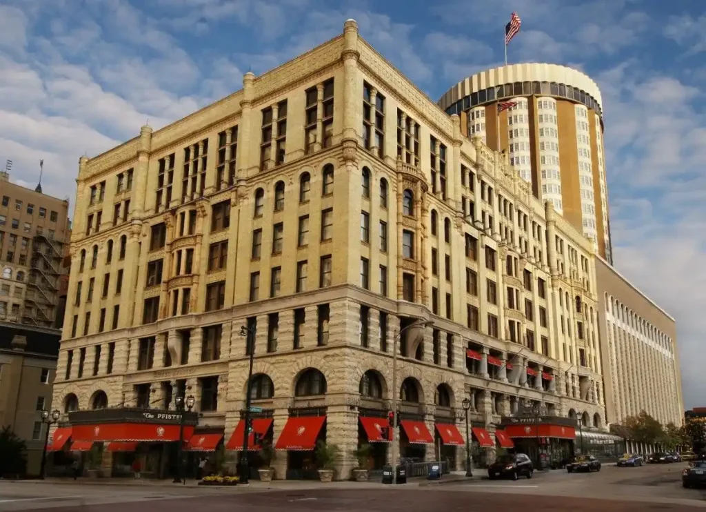 The Pfister Hotel, a historic building with a beige facade featuring arches and red awnings on the lower level, stands gracefully under a partly cloudy sky, with a modern cylindrical high-rise in the background.