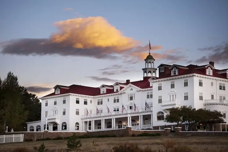 A large, white colonial-style building with a red roof, U.S. flags, and a cupola at dusk with a few clouds overhead resembles the grandeur of The Stanley Hotel.
