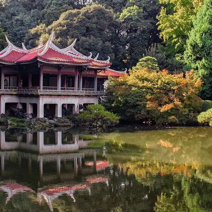 20 Bucket-List Worthy Exotic Travel Destinations to Visit Now 7 A traditional Japanese building with a red roof is reflected in a serene pond surrounded by lush greenery and autumn-colored trees. The scene is calm and picturesque, capturing the essence of nature and architecture in harmony, reminiscent of a tranquil retreat amidst the bustling life of Tokyo.