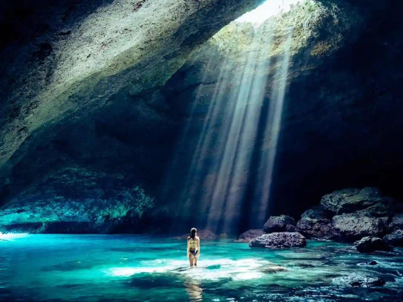 A person stands in shallow, turquoise water inside a large cave, illuminated by sunbeams streaming through a wide opening in the rocky ceiling above.